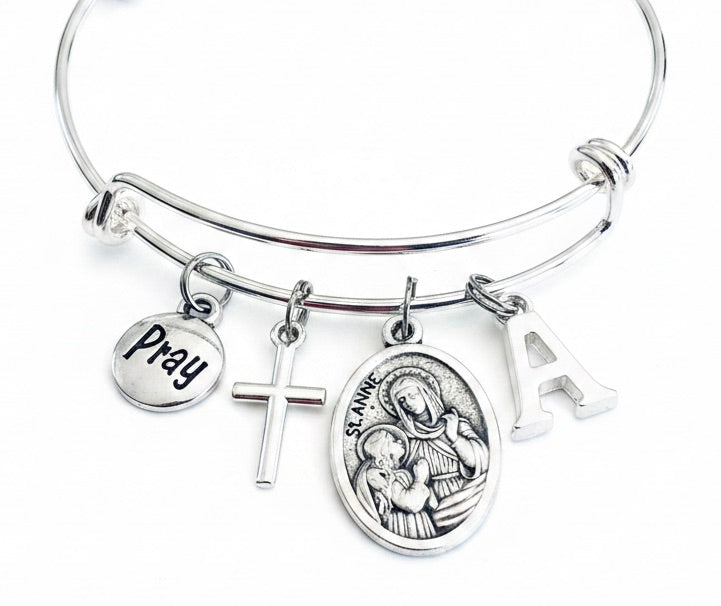 Silver expandable bangle bracelet with Saint Anne oval medal charm, silver cross, Pray disc, and personalized initial charm — Catholic jewelry gift for mothers and grandmothers by Jules Obsession