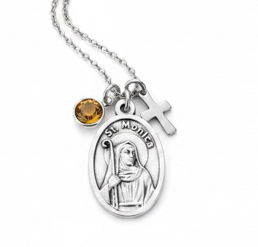 Silver St. Monica necklace featuring an oval medallion, a small cross charm, and a round amber gemstone on a white background.