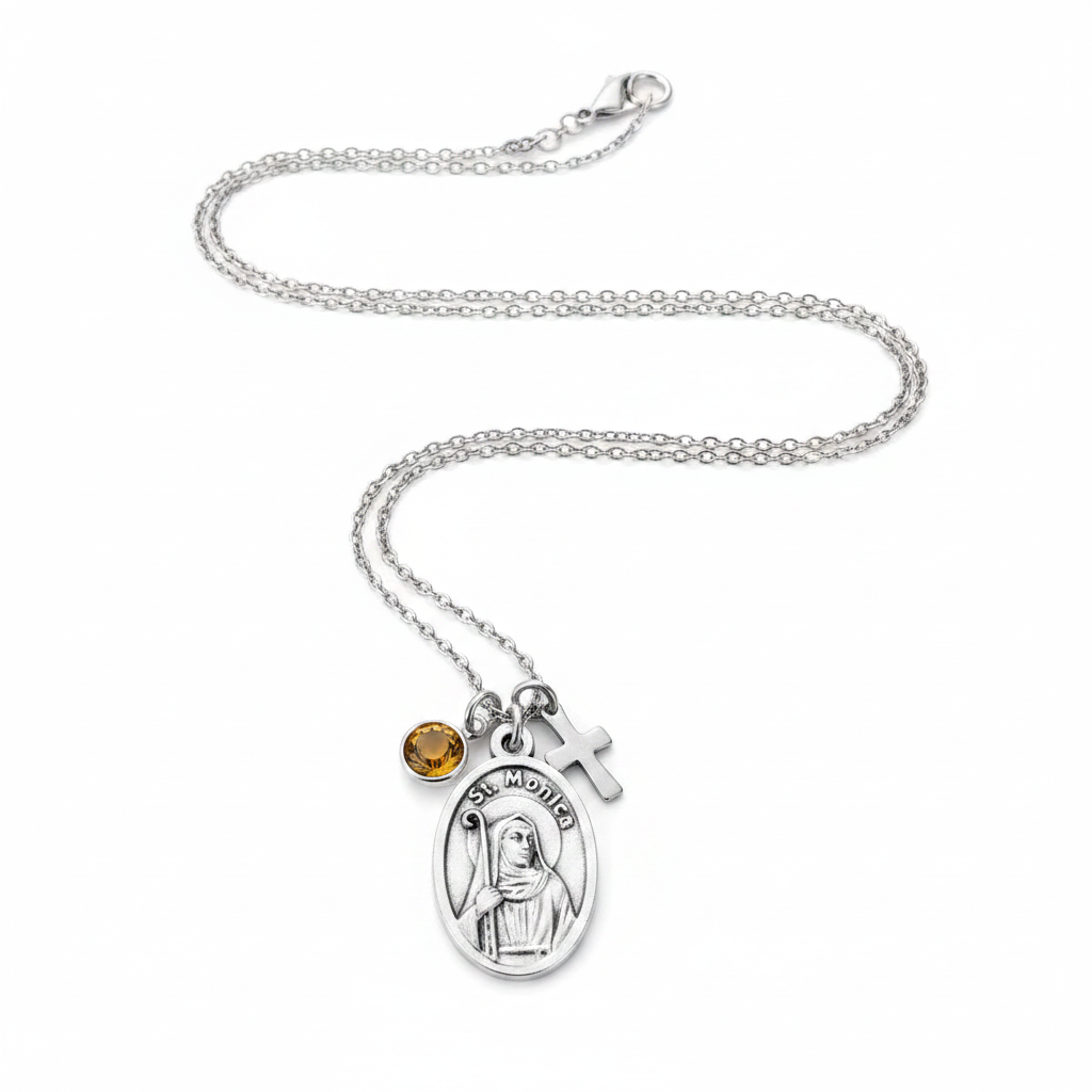 A silver chain necklace featuring an oval Saint Monica pendant, a small silver cross charm, and a round amber-yellow gemstone accent, displayed on a clean white background.