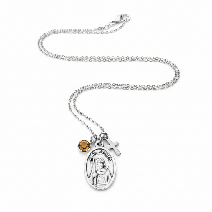 A silver chain necklace featuring an oval Saint Monica pendant, a small silver cross charm, and a round amber-yellow gemstone accent, displayed on a clean white background.