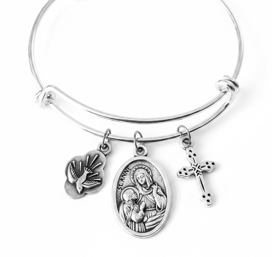 A silver-toned expandable bangle bracelet with three dangling religious charms: a dove, an oval Saint Anne medallion, and a cross, shown on a plain white background.