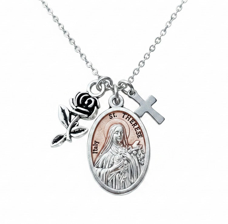Silver chain necklace on white background featuring an oval Italian St. Therese of Lisieux religious medal with rose-gold enamel center and bas-relief image of St. Therese holding flowers, a silver rose bud charm with stem and leaves, and a small silver Latin cross charm