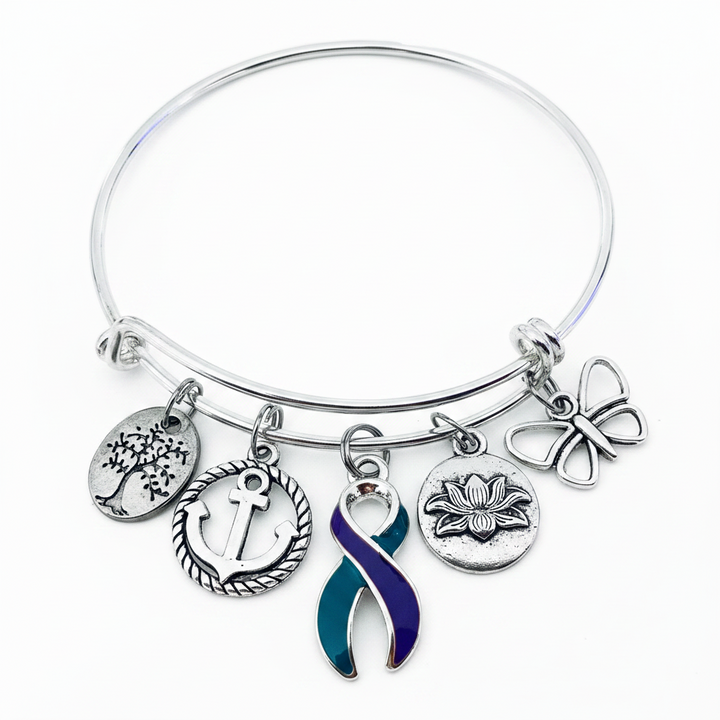 A silver-toned bangle bracelet with five dangling charms, including a tree, anchor, lotus flower, butterfly, and a teal-and-purple awareness ribbon, shown on a plain white background.
